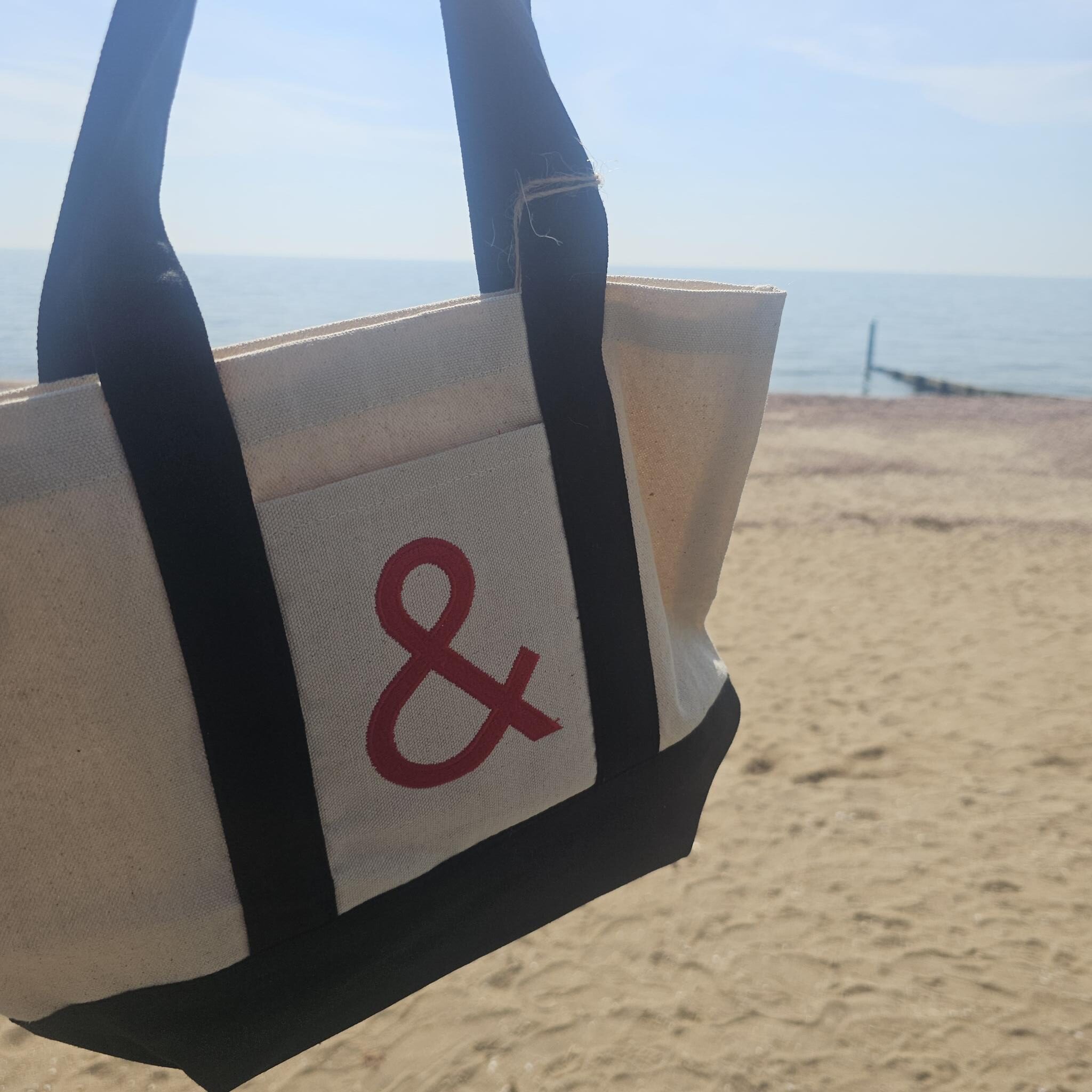A canvas tote bag on the beach with black handles and a red ampersand